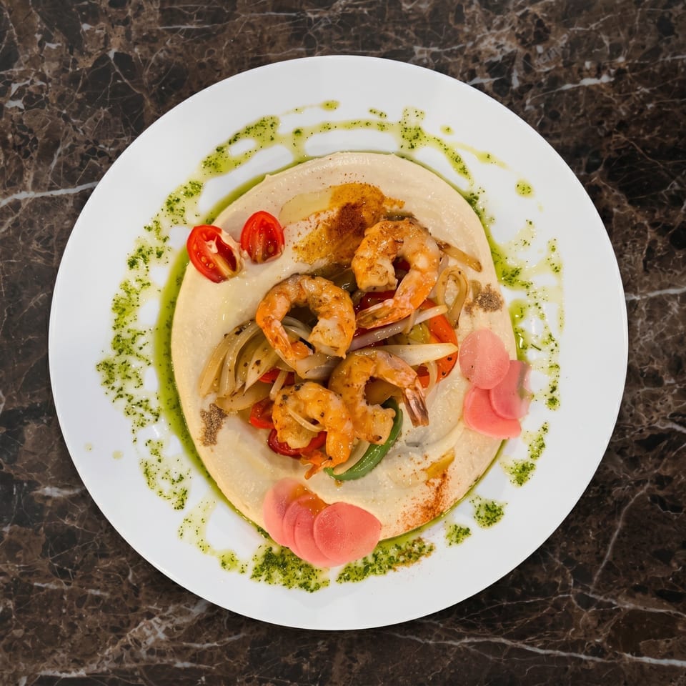 Overhead view of a white plate with hummus, sautéed shrimp, vegetables, and a green sauce.